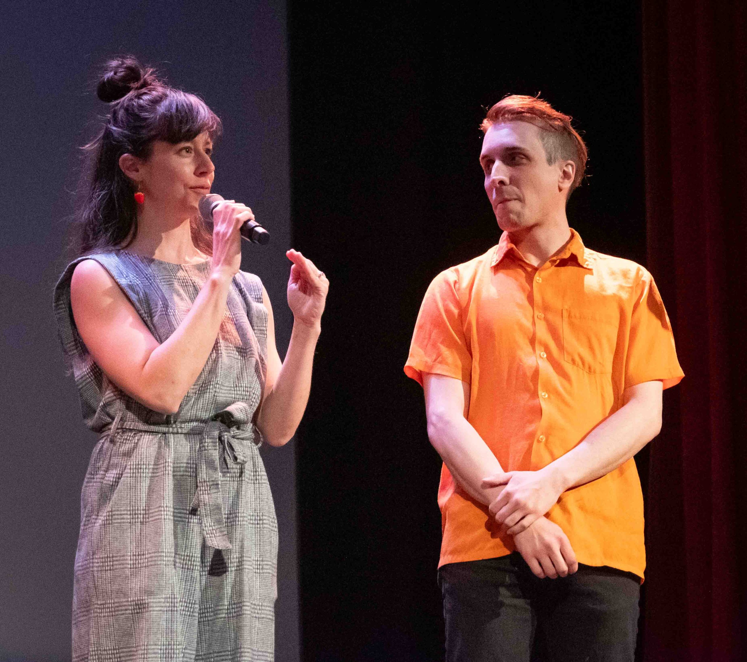 Auden Lincoln-Vogel stands on stage with Stephanie Miracle and watches as she speaks through the microphone.