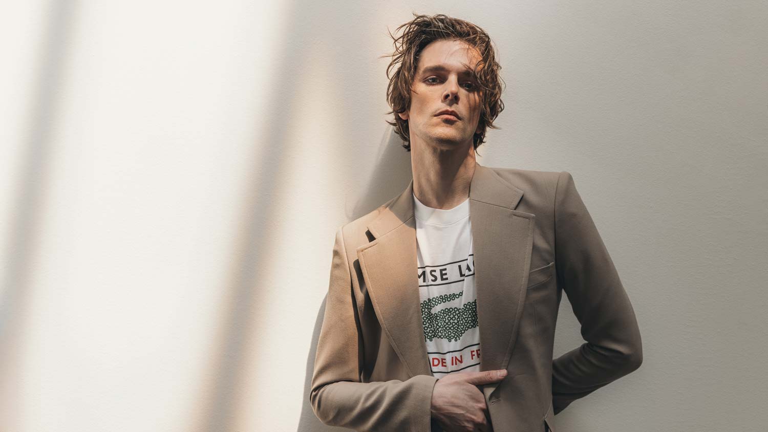 Dallon Weekes is shown from the waist-up, leaning against a white wall. They wear a tan suit jacket over a graphic T-shirt.