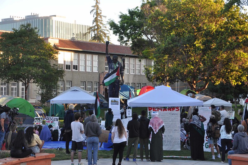 The Palestinian Flag is draped over a bronze statue looking down with a fist up in the air. Many protesters gather below.