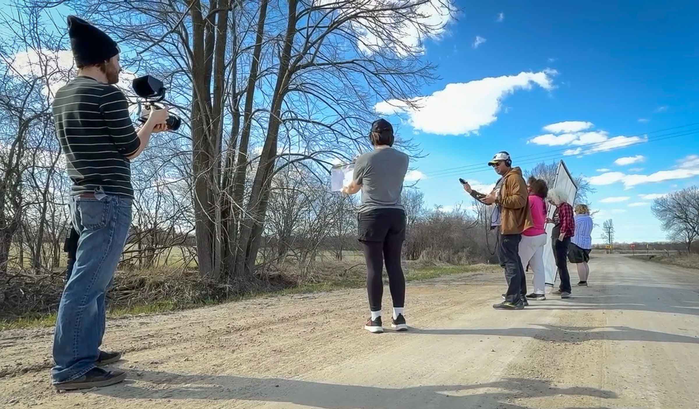 Six students stand in a line down a dirt road while filming under a bright blue sky.