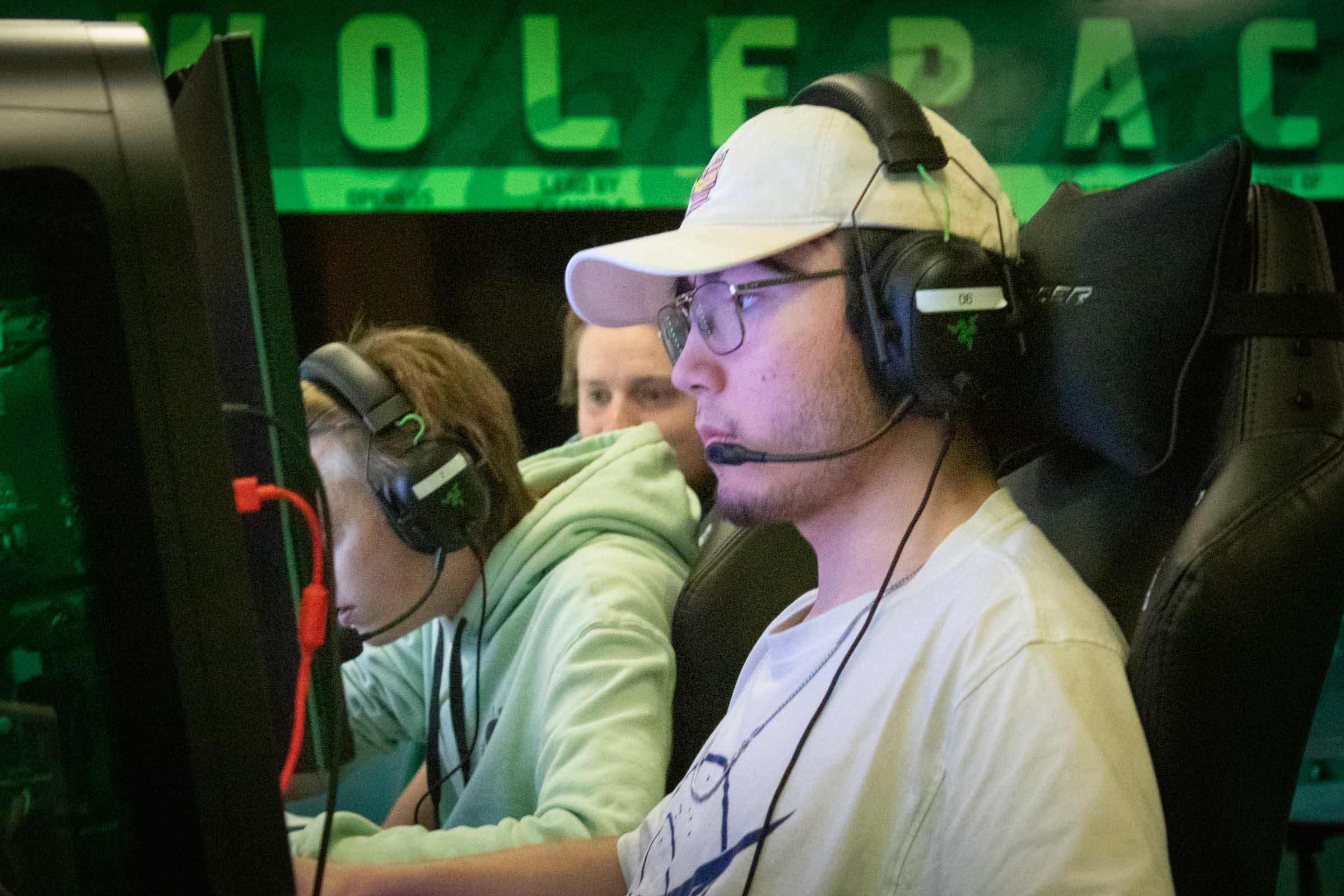 A student wears a headset and concentrates on his computer monitor. Another does the same in the background.
