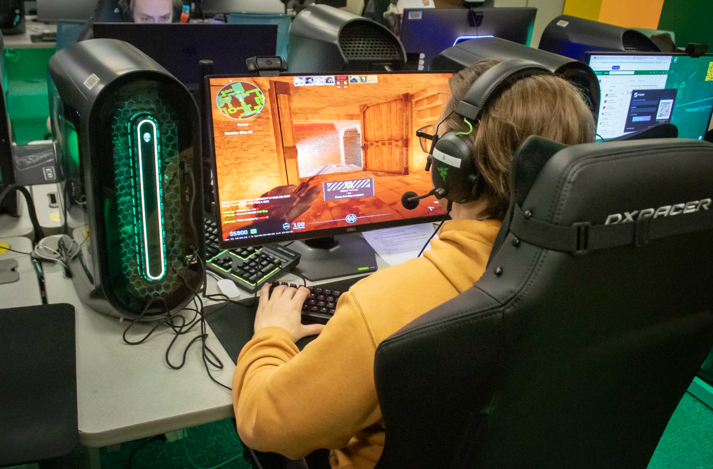 A student wears a headset and sits at a desk in front of a computer monitor while gaming.