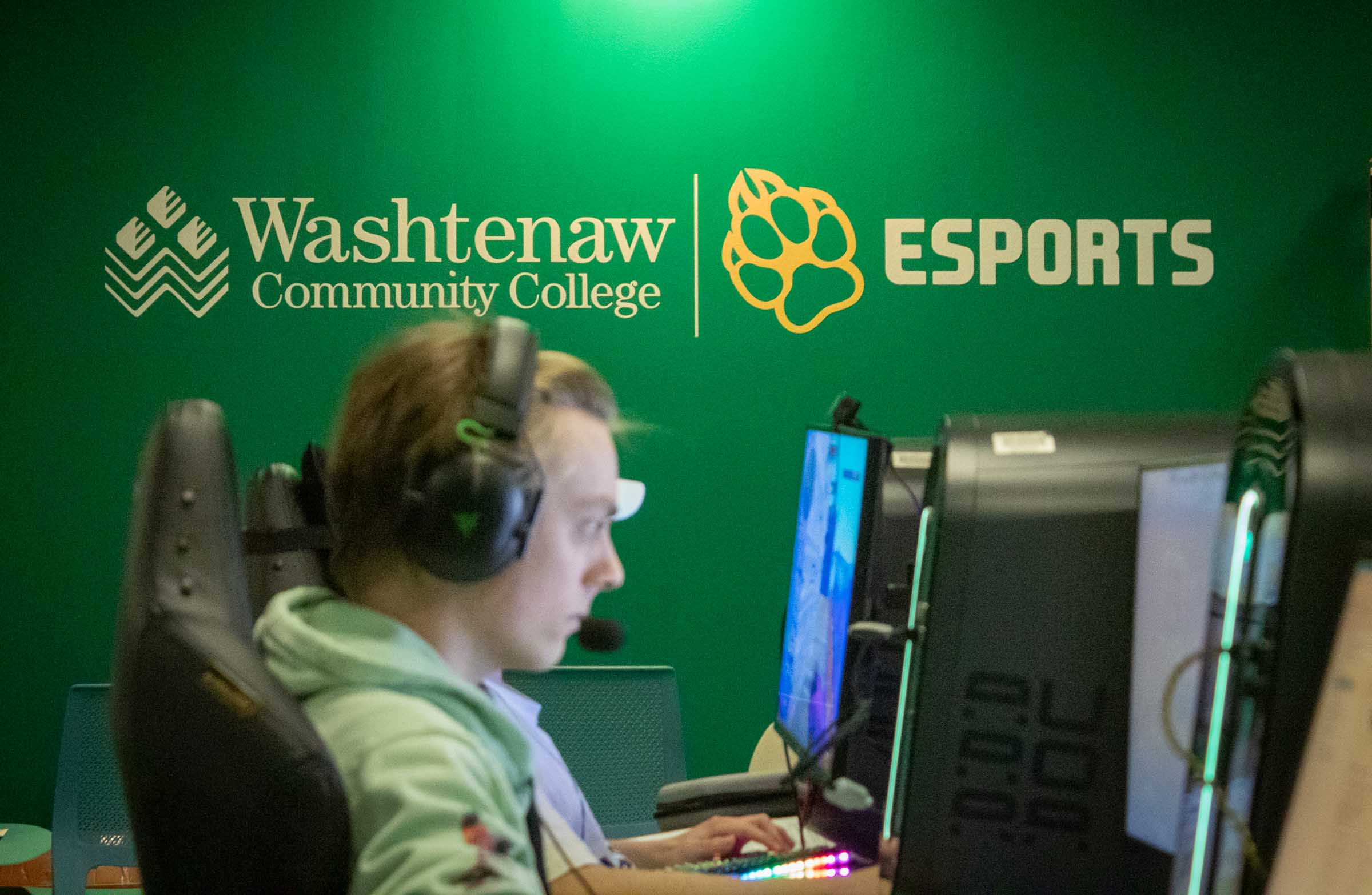 A green wall with decals reads "Washtenaw Community College Esports." An out-of-focus student games in the foreground.