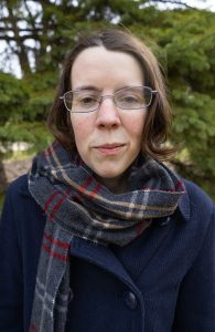Alice McGuire is a white woman with short brown hair and is standing in front of green trees.