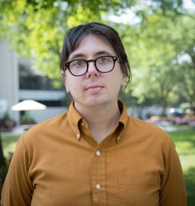 Skye is a white person with dark brown hair and they are standing in front of a green tree.