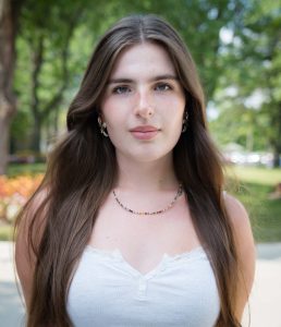 Sofia is a white woman with long brown hair and she is standing in front of a background of green trees.