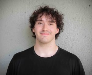 Jack is a white man with curly brown hair and he is standing in front of a gray wall.