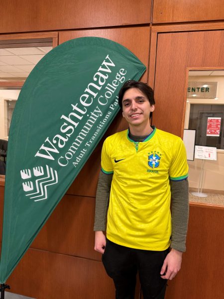 Felipe Carvalho Kalume stands in a yellow soccer jersey in front of a green Washtenaw Community College banner.