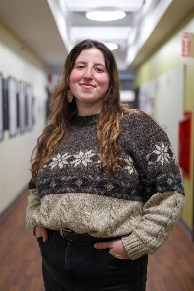Ameera Salman, a white person with long brown stands next to The Voice newsroom smiling with their hands in their pockets. They are wearing black pants and a brown sweater.