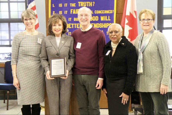 Five people stand inside, posing with a plaque in front of the Rotary Club backdrop and an American and Canadian flag.